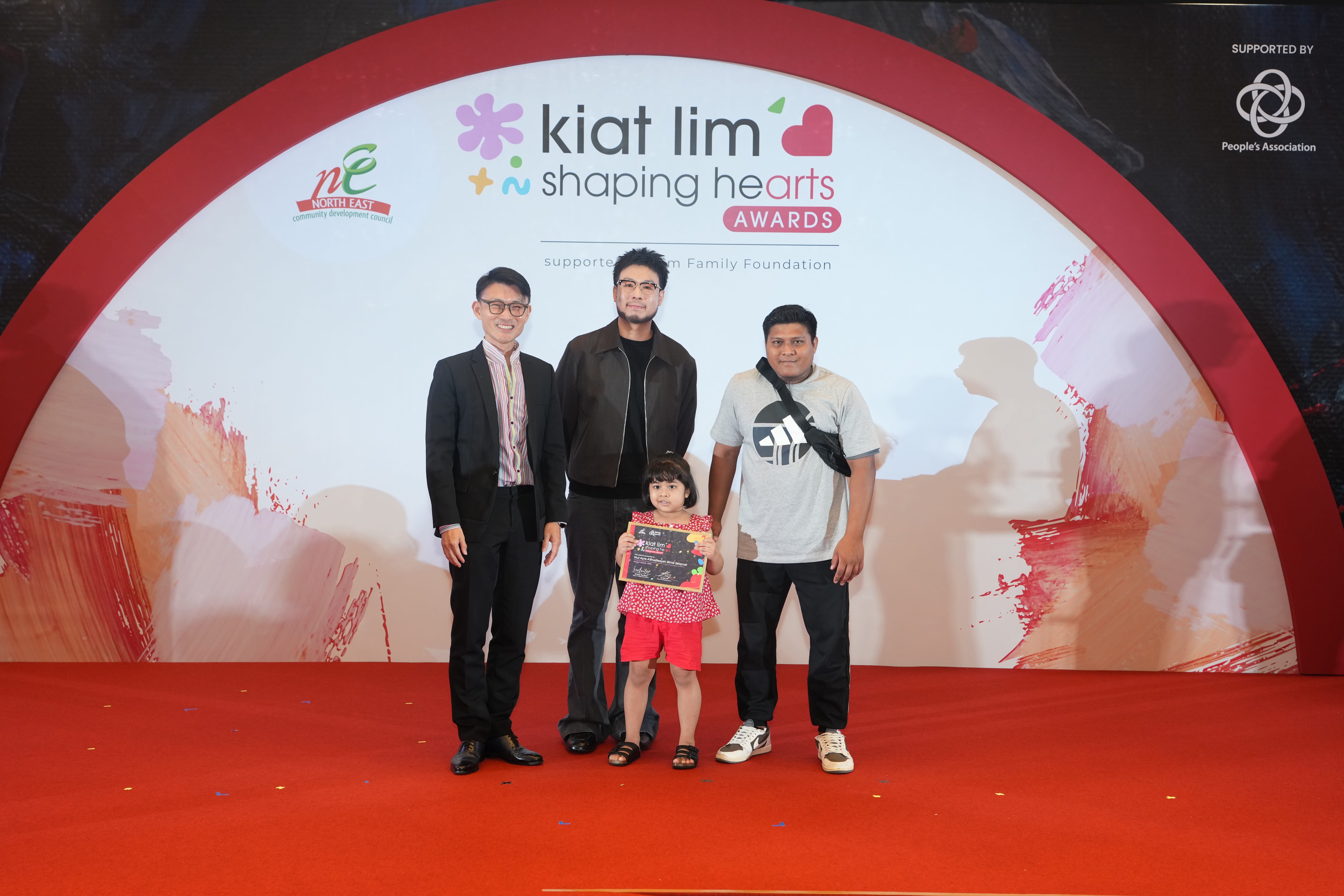Award recipient standing on stage with the presenter, holding their certificate and posing for a group photo during the award ceremony, with a backdrop reading ‘Kiat Lim-Shaping Hearts Awards'.